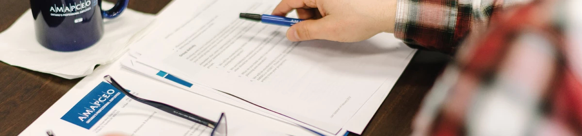 Hands of a member are placed over a number of AMAPCEO-branded papers. They are resting their glasses down with one hand and holding a pen with another.