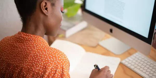 A person working from home, writing in a notebook and in front of a computer.