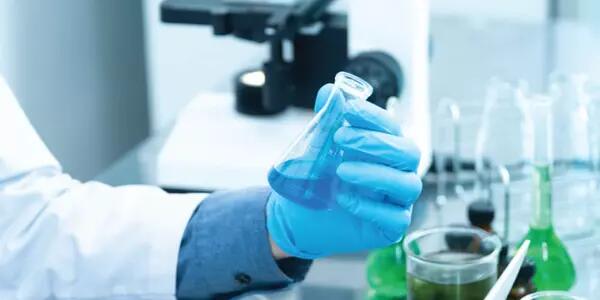 A hand in a blue latex glove is holding a small beaker with a blue liquid. The person is wearing a lab coat and it is in a laboratory setting.