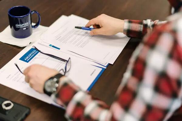 A person's hands holding eyeglasses and a pen, with an AMAPCEO mug and AMAPCEO files on the table in front of them