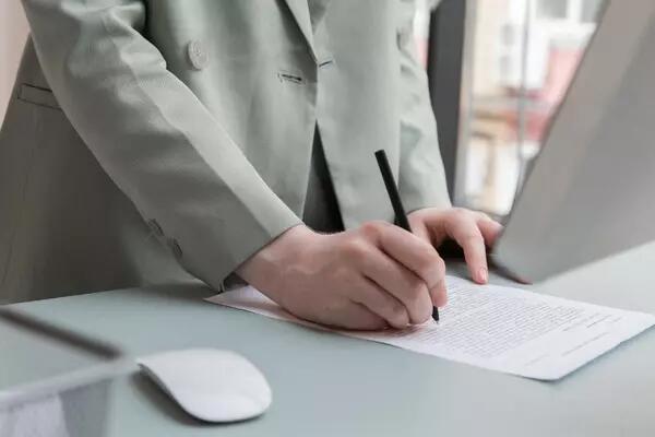 A person's hands taking notes on a paper with a monitor and mouse on the desk
