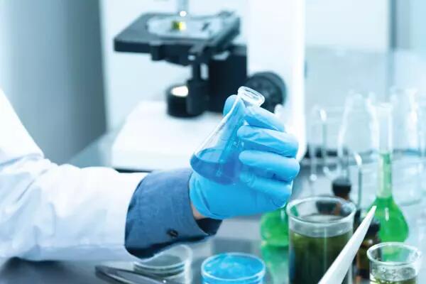 A hand in a blue latex glove is holding a small beaker with a blue liquid. The person is wearing a lab coat and it is in a laboratory setting.