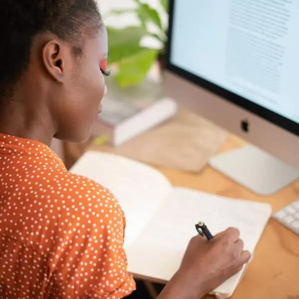 A person working from home, writing in a notebook and in front of a computer.